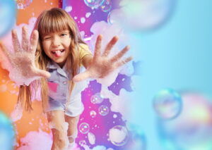 Joyful girl with glitter on her face reaching toward the camera amid colorful bubbles and a rainbow background.