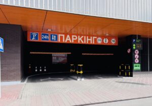Entrance to a parking garage with an orange canopy and bold red Ukrainian 'ПАРКІНГ' sign, showing 24h parking and bike and height/weight restrictions outside.