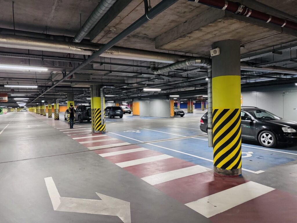 Underground parking garage with yellow striped columns, crosswalk markings, and several parked cars; a person walks toward a vehicle.