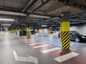 Underground parking garage with yellow striped columns, crosswalk markings, and several parked cars; a person walks toward a vehicle.