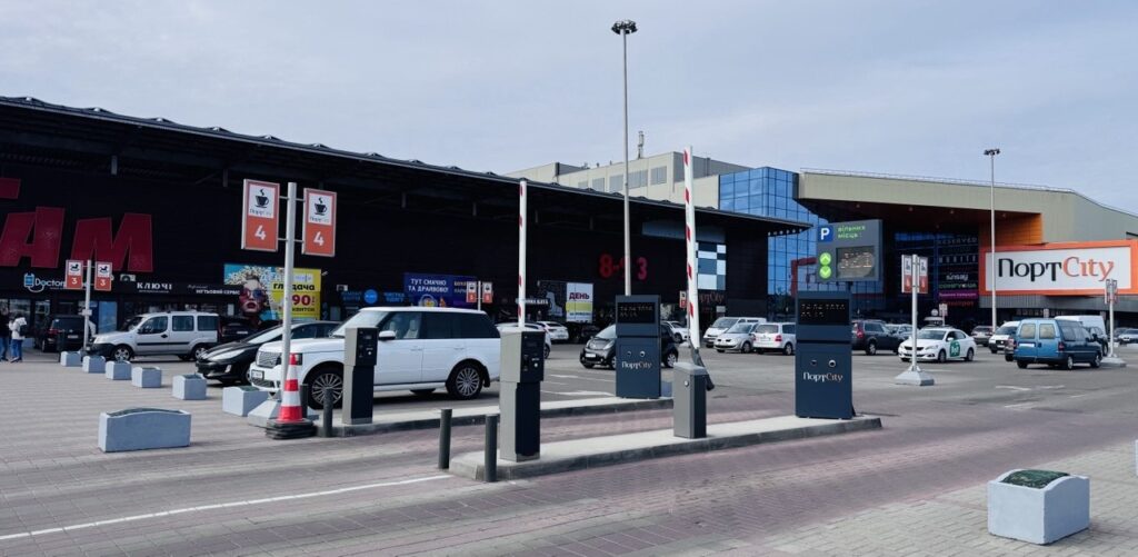 PortCity shopping center parking lot with cars, barrier gates, and payment kiosks in front of a large building.