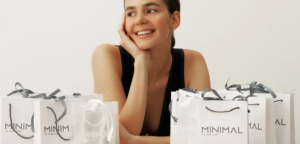 Woman smiling while resting her chin on her hand, surrounded by white gift bags labeled MAXIMAL or MINIMAL UA Jewellery on a light backdrop